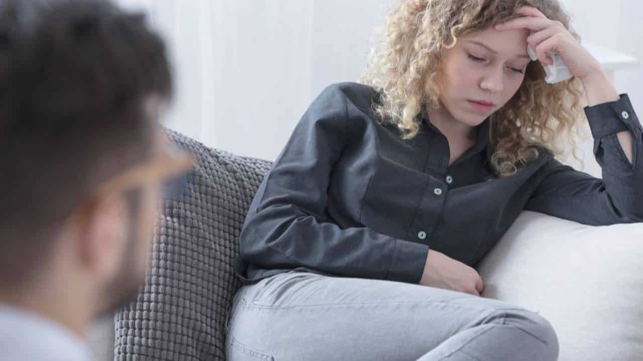 Woman with curly hair resting her head on her hand on a sofa, with a man in glasses seen from behind in the foreground