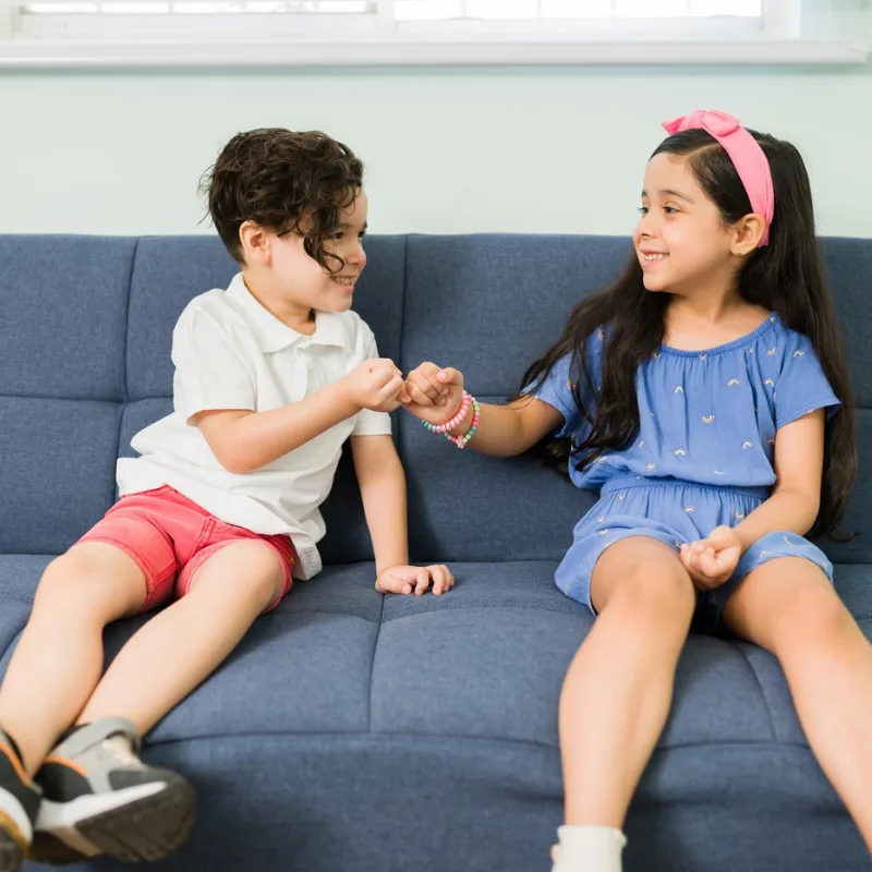 Boy and girl sitting side by side on a blue sofa, bumping fists and smiling