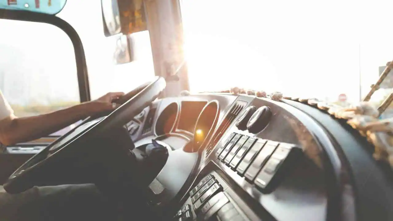 Close-up view of a truck driver's hand on the steering wheel inside a sunlit truck cabin, with dashboard controls and sunlight streaming through the window.