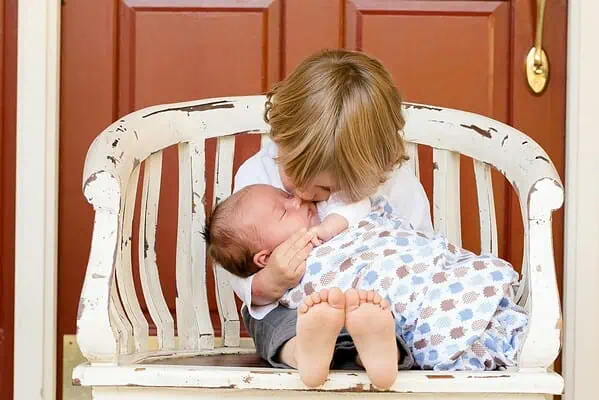 Older brother gently hugging and kissing his baby sibling while sitting on a white wooden bench.