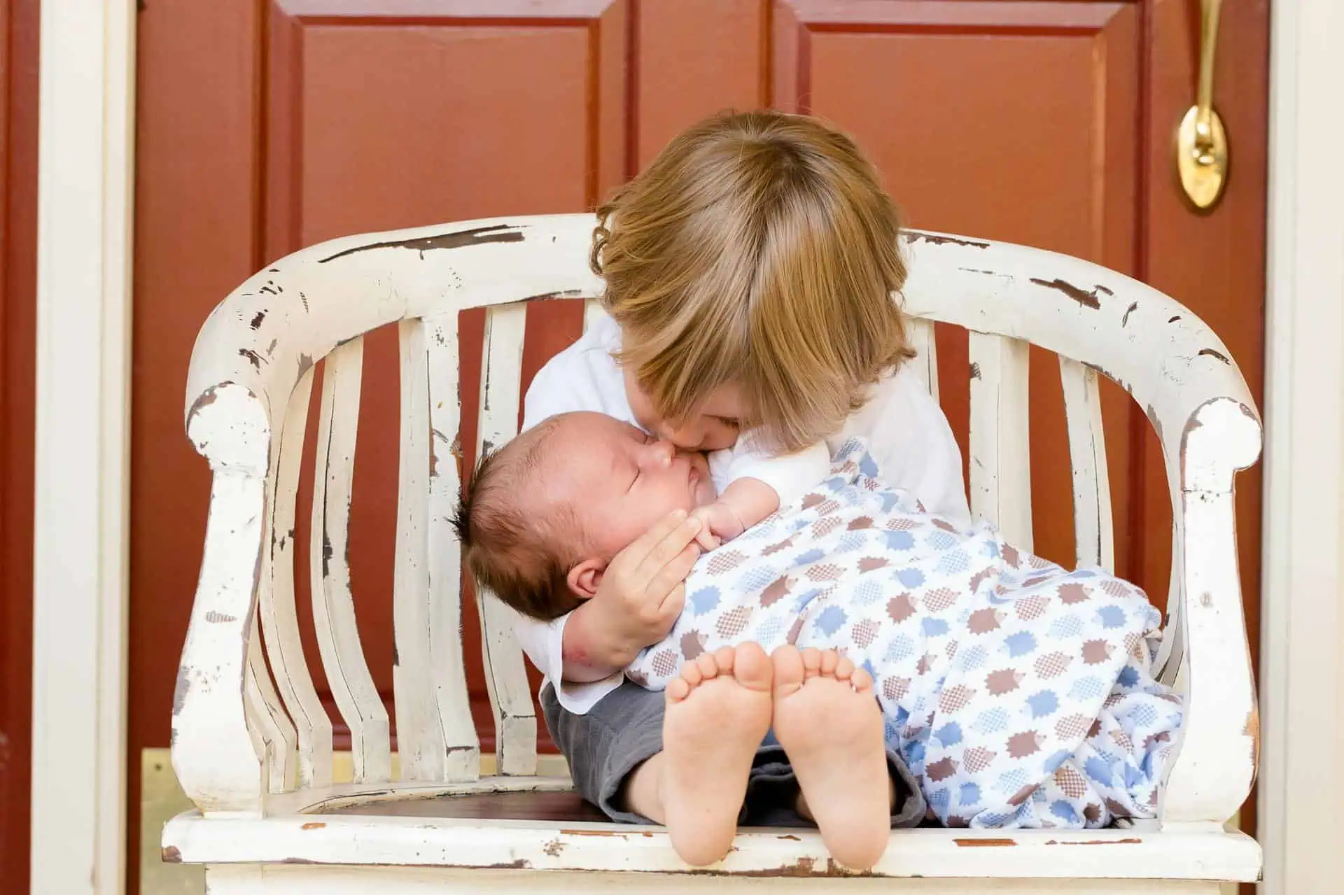 Older brother gently hugging and kissing his baby sibling while sitting on a white wooden bench.
