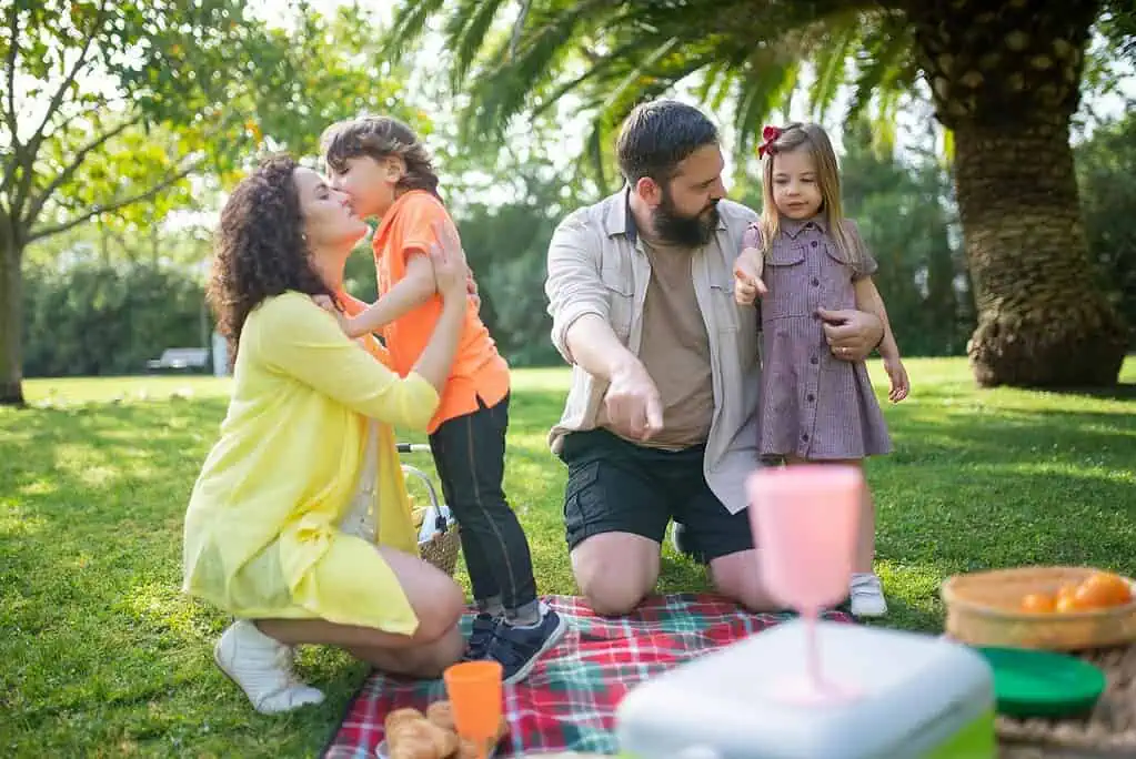 Family enjoying a picnic outdoors, with a mother hugging her son and a father talking to his daughter on a blanket.