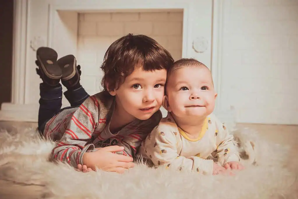 Older brother lying on a soft rug beside his baby sibling, both looking forward with warm expressions.