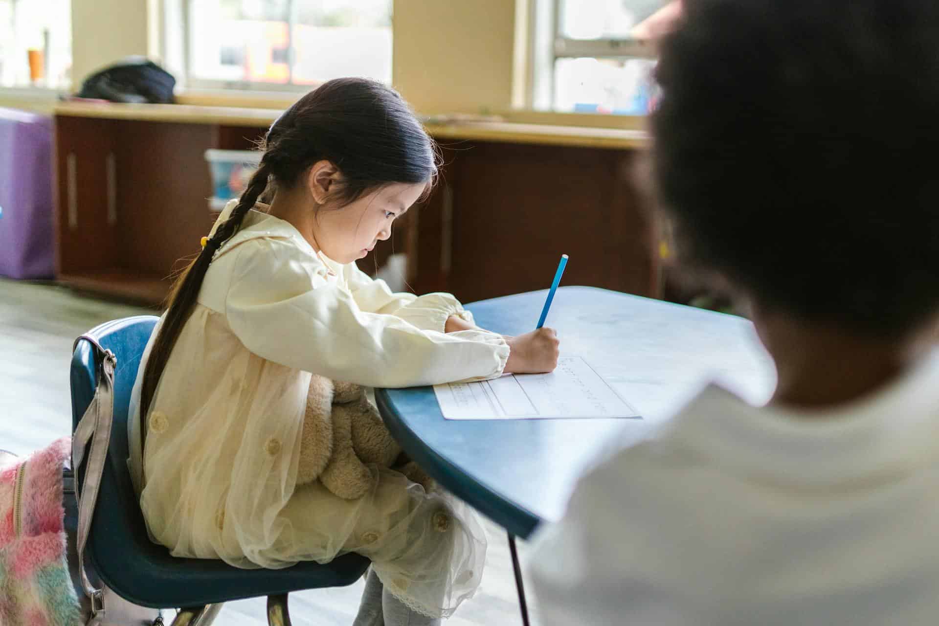Young girl in a classroom writing on a worksheet with a blue pencil, a soft toy on her lap and another child sitting opposite.