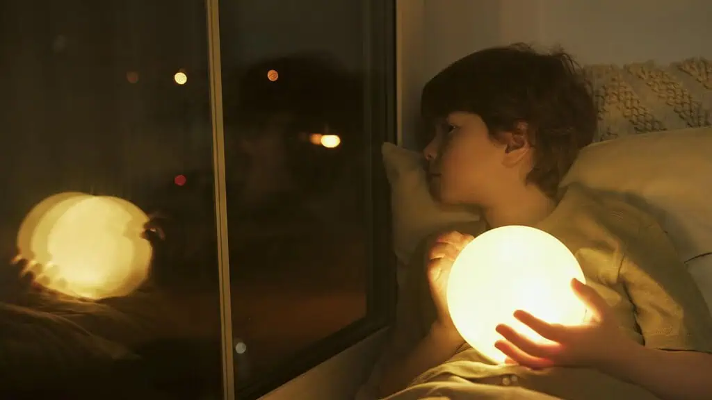 Young boy lying in bed at night holding a glowing light, staring out the window with a restless expression