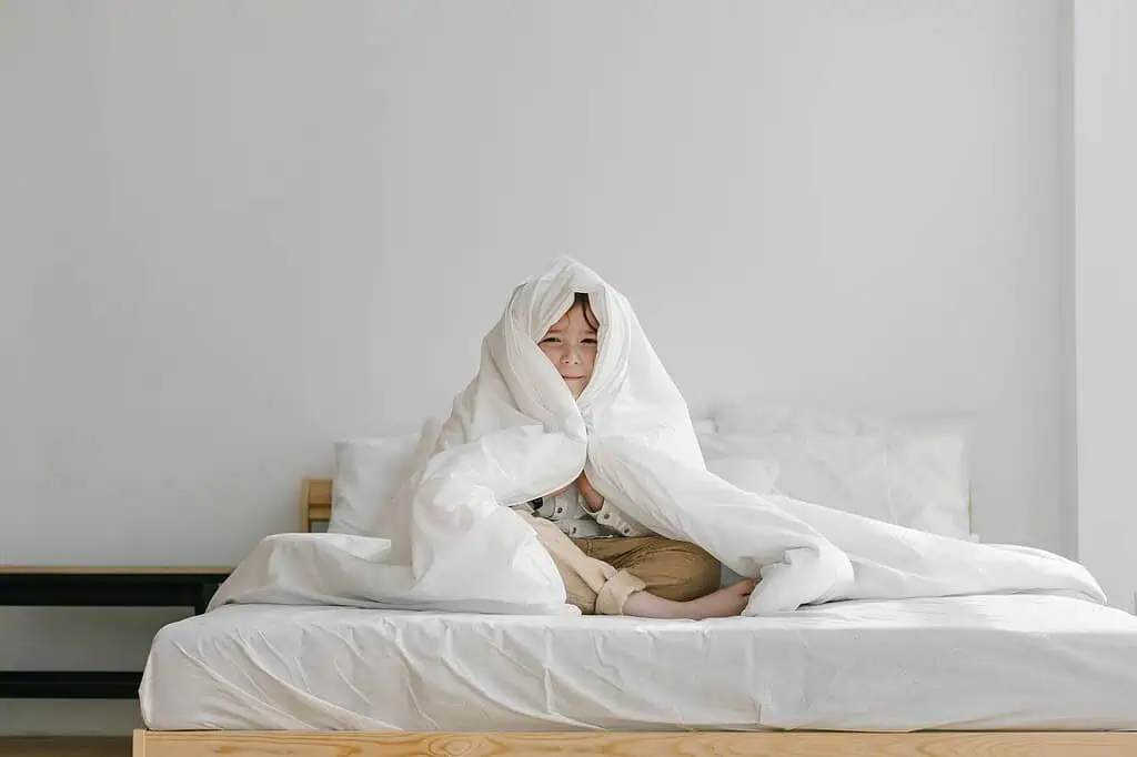Young boy sitting in bed wrapped in a white blanket, showing signs of sleep trouble or restlessness