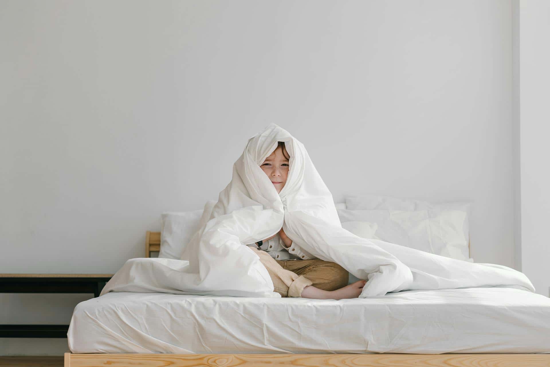 Young boy sitting in bed wrapped in a white blanket, showing signs of sleep trouble or restlessness