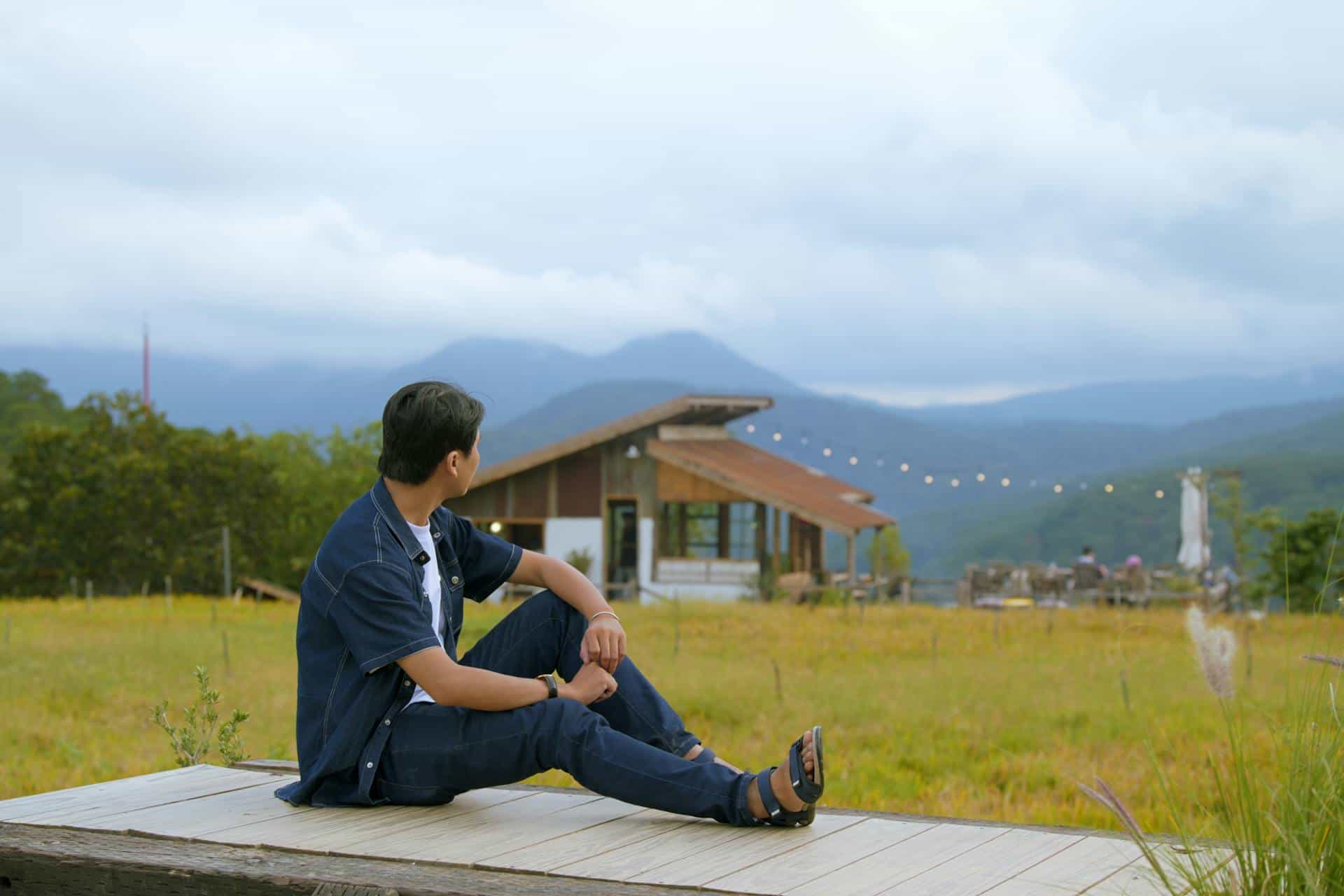 Adult male sitting alone outdoors, looking toward a cabin in the distance with mountains in the background representing quiet reflection and mental escape often needed by adults with ADHD.