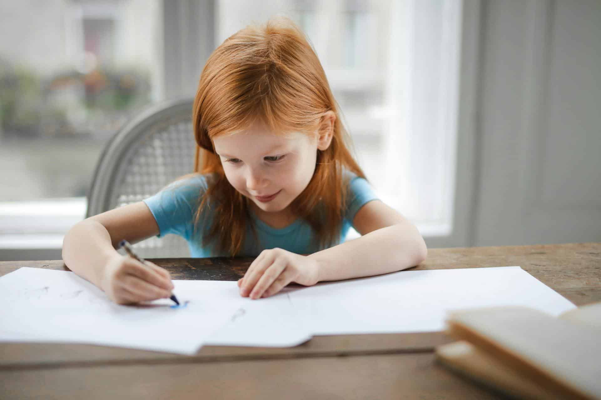 Young red-haired girl smiling as she writes on paper at a wooden table by a window.
