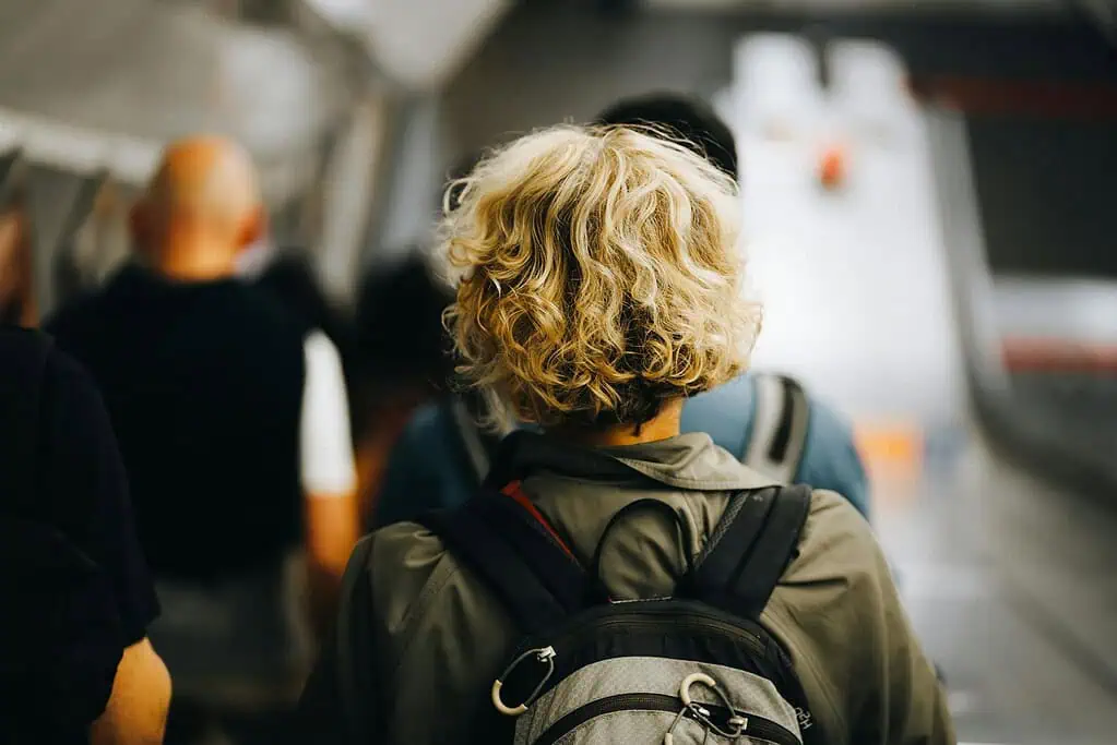 Back view of an adult with curly blonde hair wearing a backpack while walking through a busy station, symbolizing the daily challenges of dyspraxia in adults.