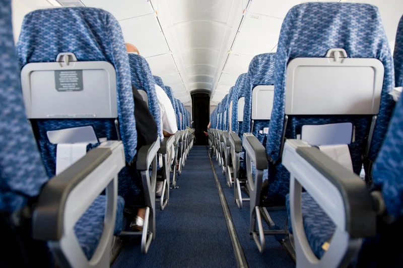 An interior view of an airplane cabin taken from the aisle, showing rows of blue patterned seats with tray tables and passengers seated, capturing a typical in-flight experience.