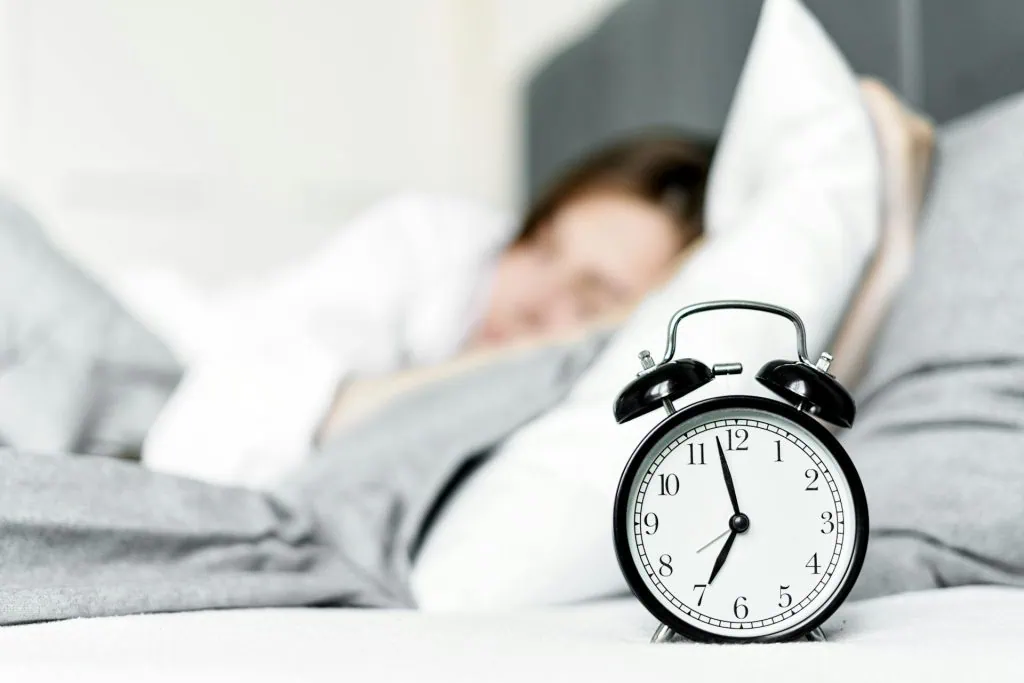 Close-up of an alarm clock on a bed with a person sleeping in the background, highlighting sleep time management