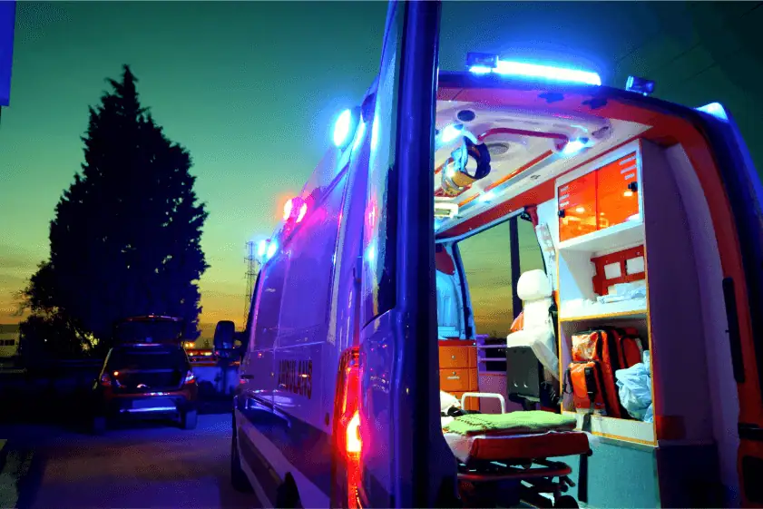 An ambulance with its rear doors open is illuminated by bright emergency lights during twilight. The interior is stocked with medical equipment, including a stretcher, oxygen tanks, and first aid supplies. A parked car and a darkening sky with power lines are visible in the background.