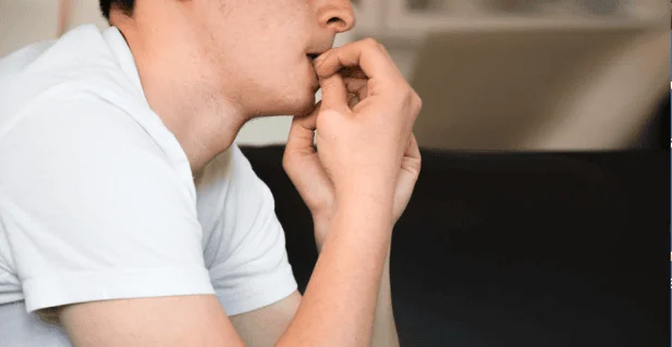 A young man in a white t-shirt sitting on a couch, nervously biting his nails while appearing deep in thought or worried about something.