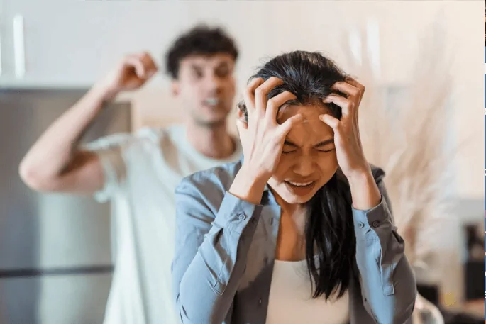 A distressed woman holding her head while a man in the background is angrily gesturing, depicting a tense argument or domestic conflict.