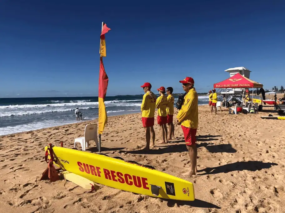 Lifeguards in yellow and red uniforms standing on a sandy beach near the shoreline with a surf rescue board.