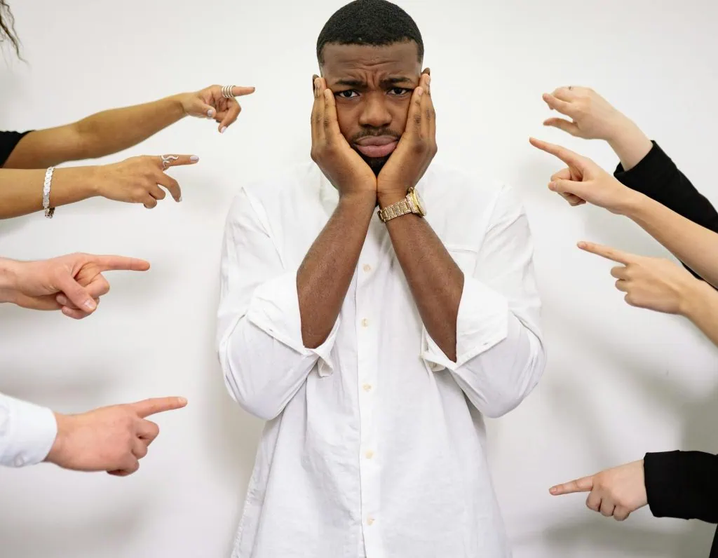 Worried man standing while multiple hands point fingers at him in accusation or blame.