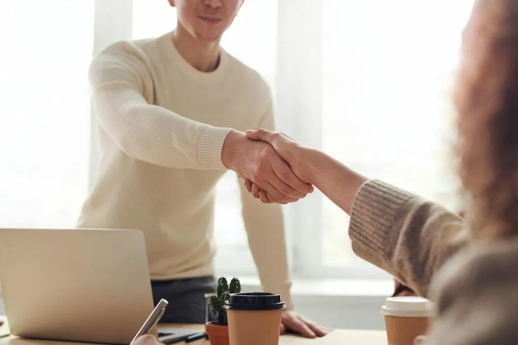 Two people shaking hands across a desk in a bright office setting, symbolizing agreement or successful partnership.