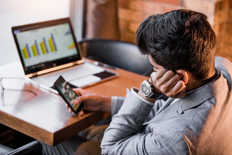 A man in a suit distracted by his phone while a laptop with a bar chart is open in front of him.