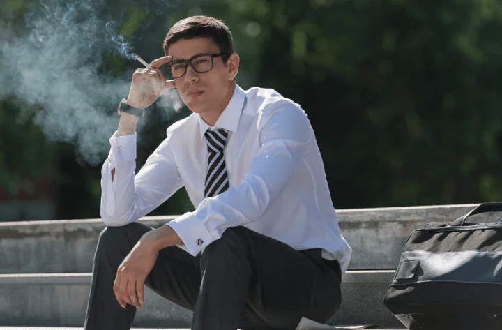 A professional man wearing a suit and glasses smokes a cigarette while sitting on outdoor steps, appearing deep in thought.