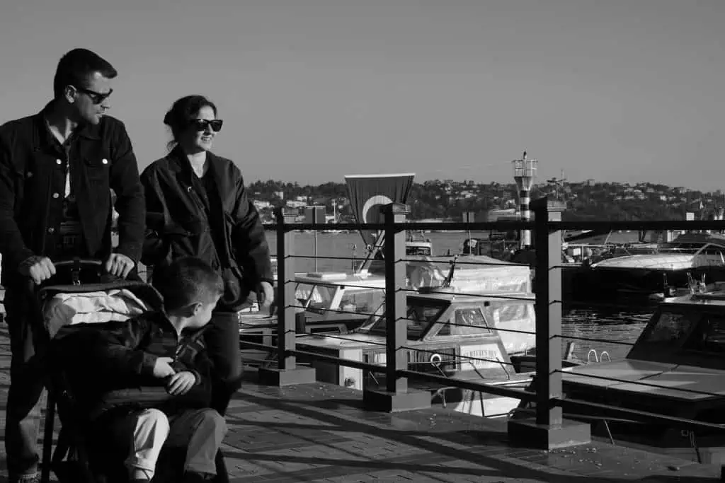 A man and woman walking together by the marina with a child in a stroller, suggesting a co-parenting moment during a family outing