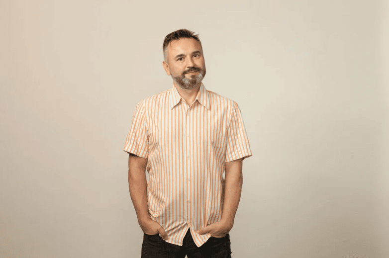 A middle-aged man with a short beard, wearing a casual orange-striped short-sleeve shirt, standing against a neutral beige background with a relaxed expression.