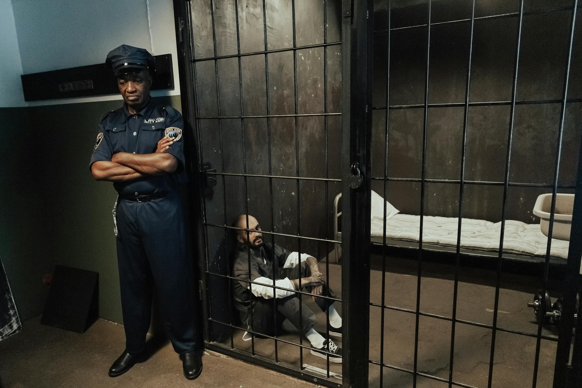 Correctional officer standing outside a prison cell with an inmate sitting on the floor, highlighting the emotional toll of the job.