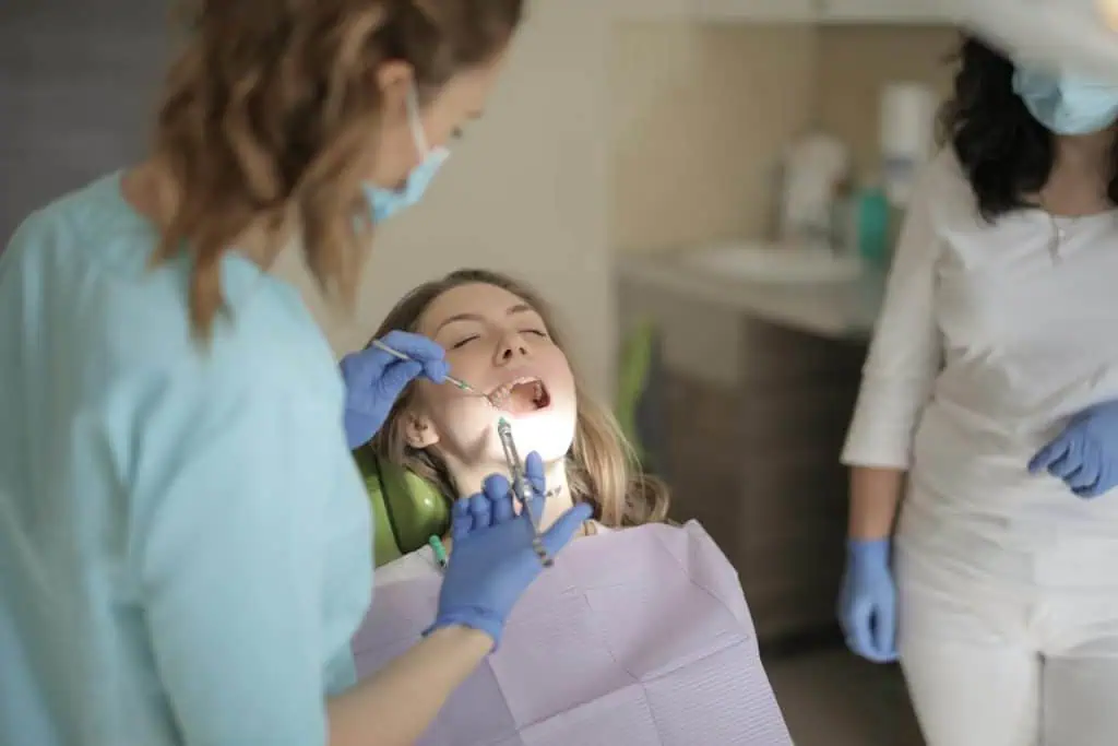 Female dentist examining a young woman’s open mouth during a dental checkup, with an assistant observing in the background.
