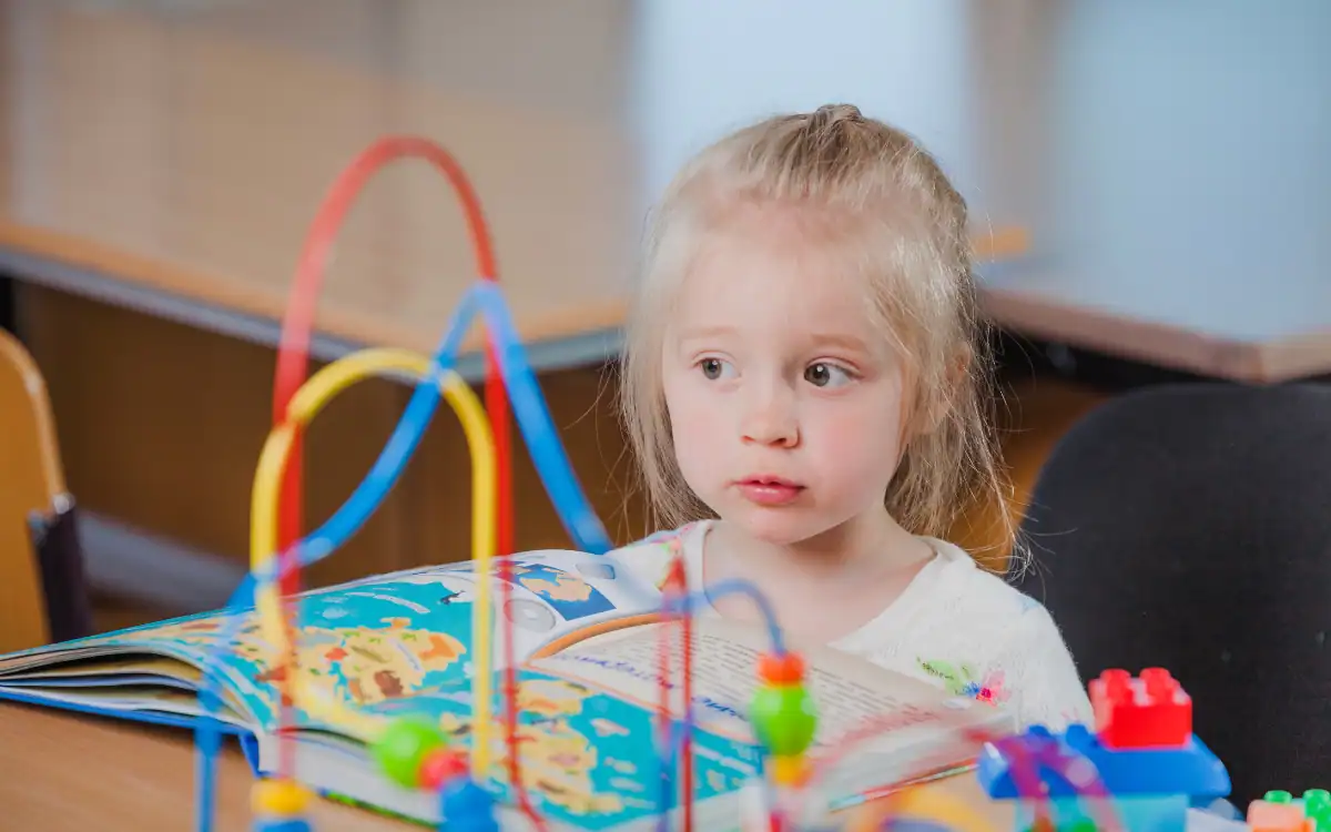Young girl with blonde hair looking to one side beside a colourful bead maze toy and a picture book