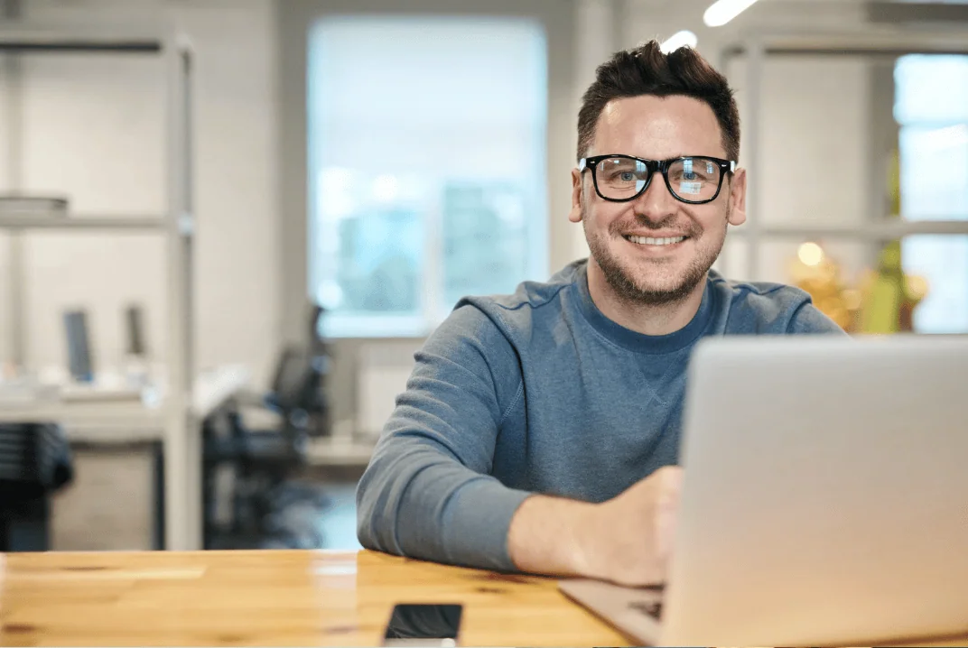 A man in a blue sweater wearing glasses, smiling while working on a laptop at a wooden desk in a modern office setting.