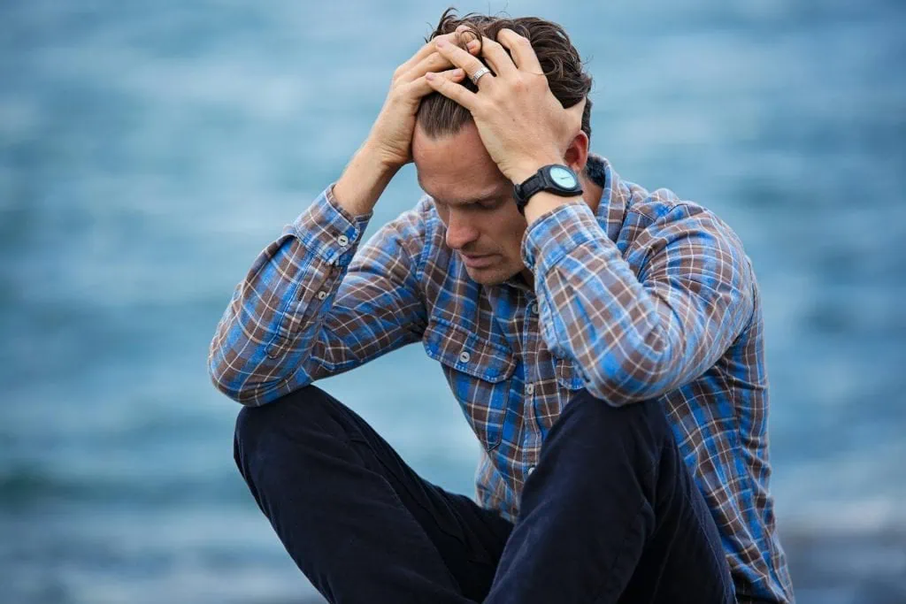 Distressed man in a plaid shirt sitting by the water, holding his head in frustration or emotional pain, possibly experiencing anxiety or PTSD.