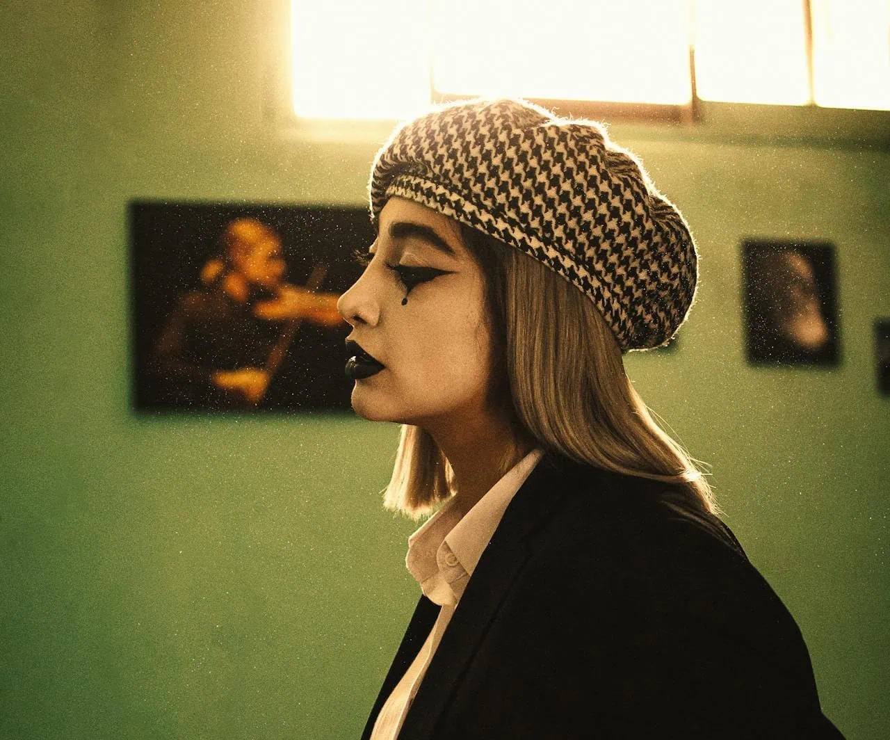 A woman with dramatic makeup, including black lipstick and teardrop eyeliner, wears a houndstooth beret and black blazer over a white shirt. She is standing in profile under warm lighting in a room with green walls and framed photos in the background.