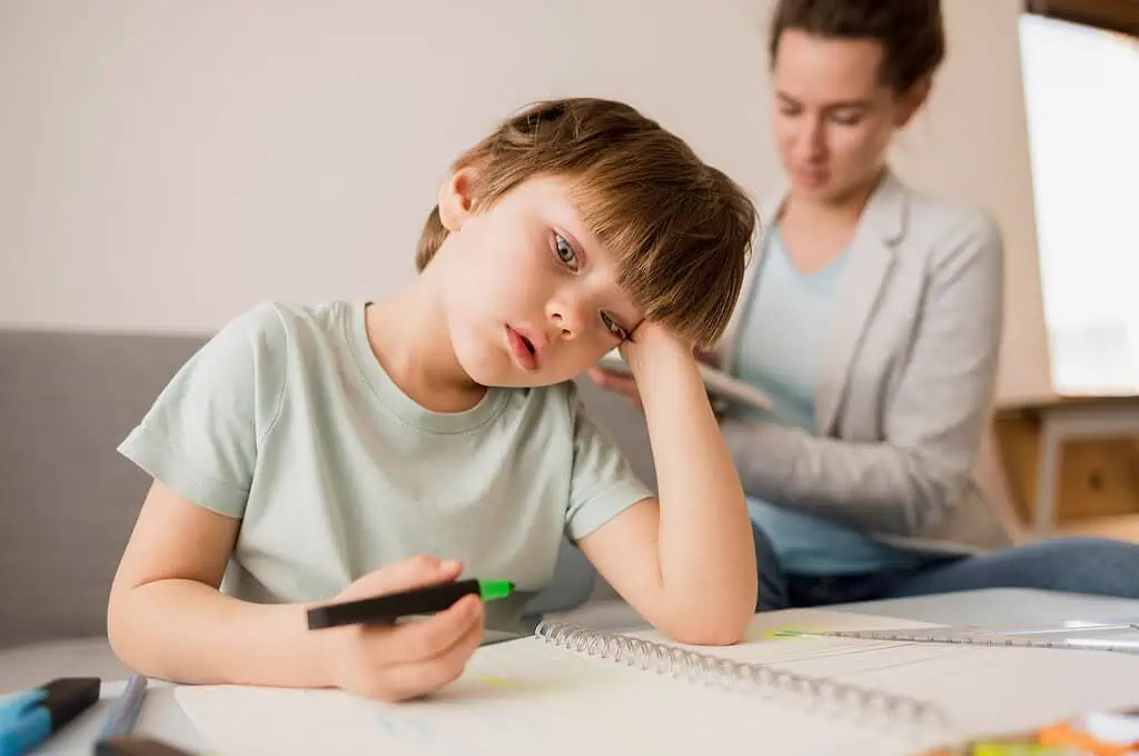 A young boy sitting at a table with a notebook, holding a marker and looking tired while an adult supports him in the background, symbolizing challenges of Dyspraxia in learning.