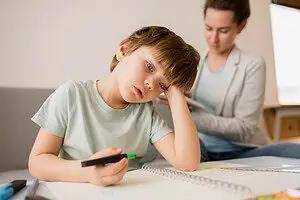A young boy sitting at a table with a notebook, holding a marker and looking tired while an adult supports him in the background, symbolizing challenges of Dyspraxia in learning.