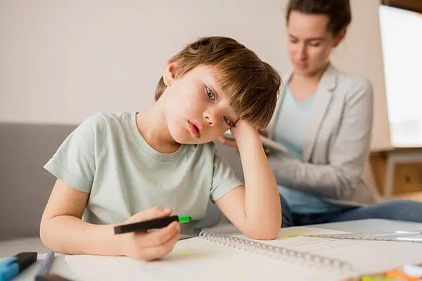 A young boy sitting at a table with a notebook, holding a marker and looking tired while an adult supports him in the background, symbolizing challenges of Dyspraxia in learning.