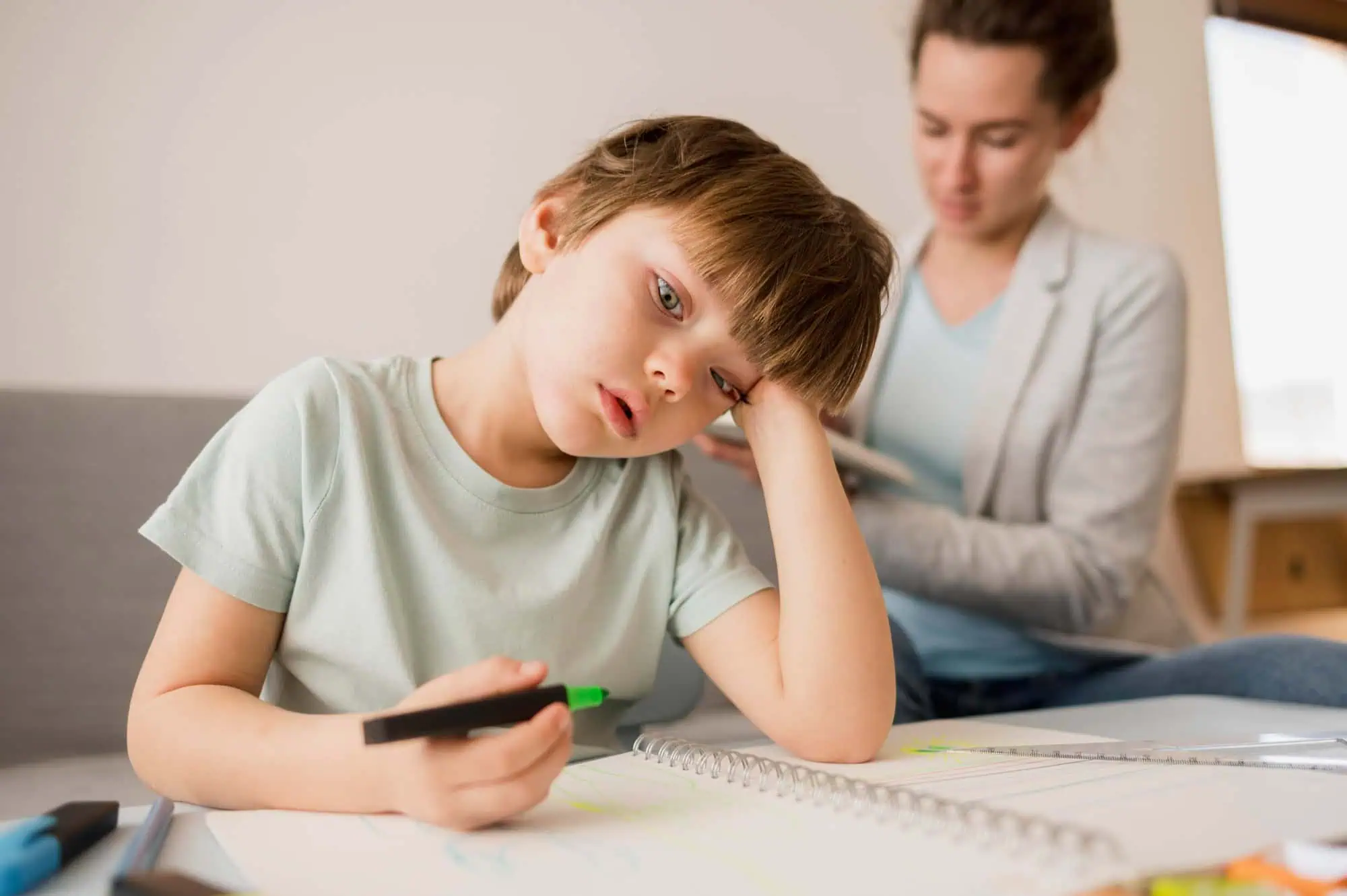 A young boy sitting at a table with a notebook, holding a marker and looking tired while an adult supports him in the background, symbolizing challenges of Dyspraxia in learning.