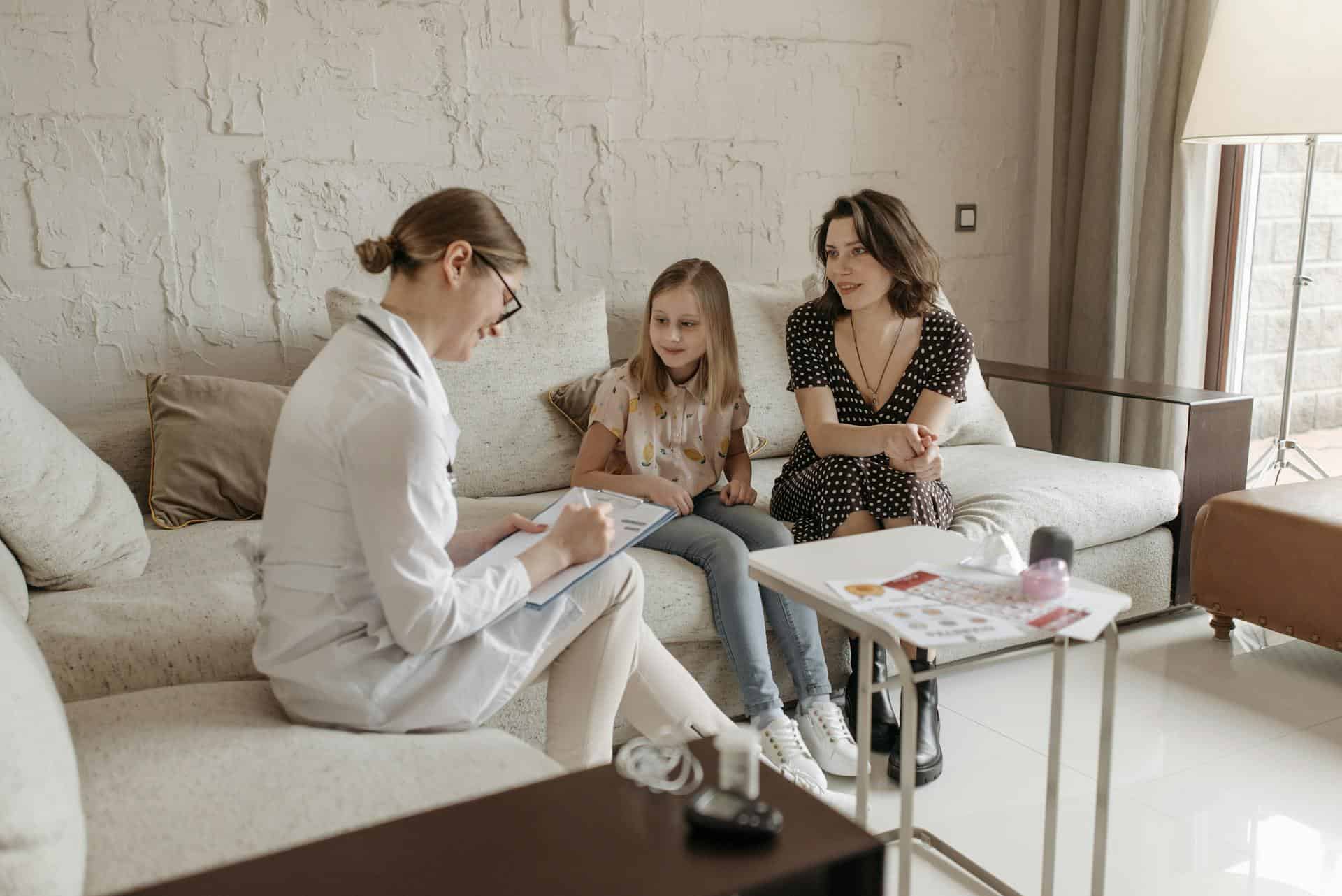 A girl and her mother sit together on a sofa while a woman in a white coat and glasses writes notes on a clipboard.