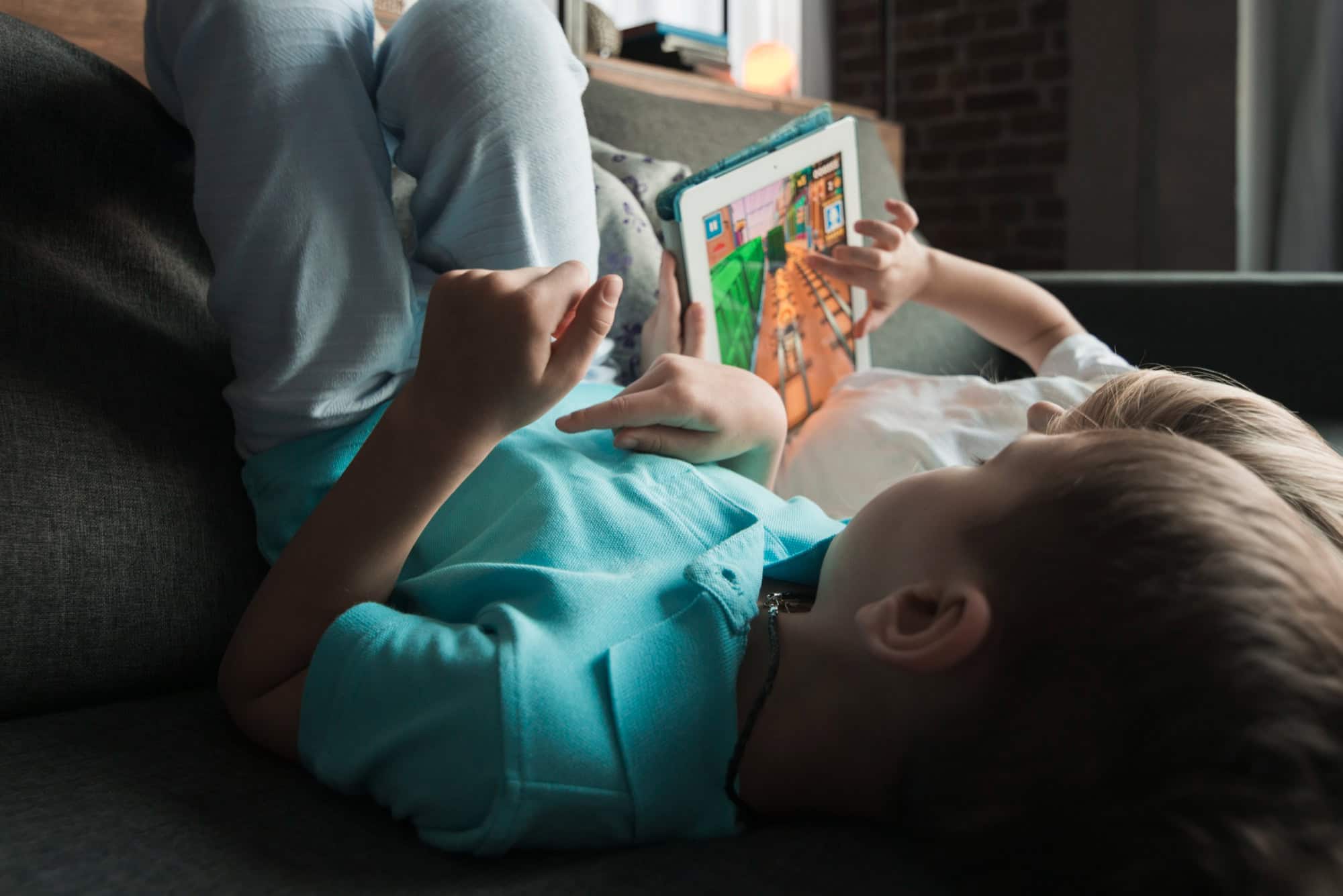 Two children lying on a sofa and playing a game on a tablet.