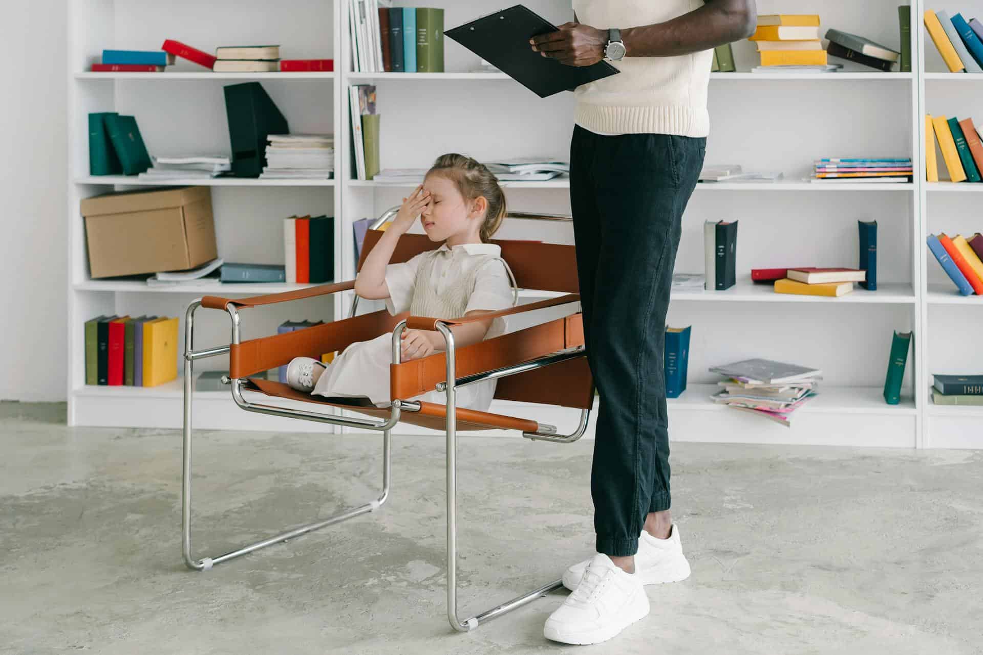 A girl sitting in a chair with her hand pressed to her forehead and her eyes shut, while an adult stands beside her holding a clipboard.