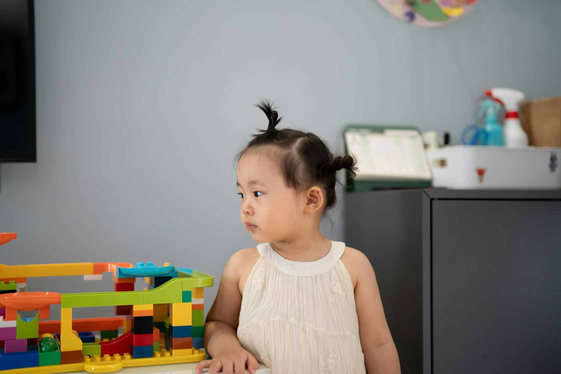 A young girl with two small buns in her hair looking off to one side, standing beside a colourful marble run built from toy building blocks.
