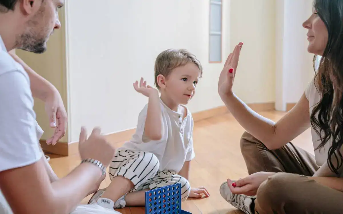 Adult in a white t-shirt and a woman giving a toddler a high five while the toddler plays with building blocks