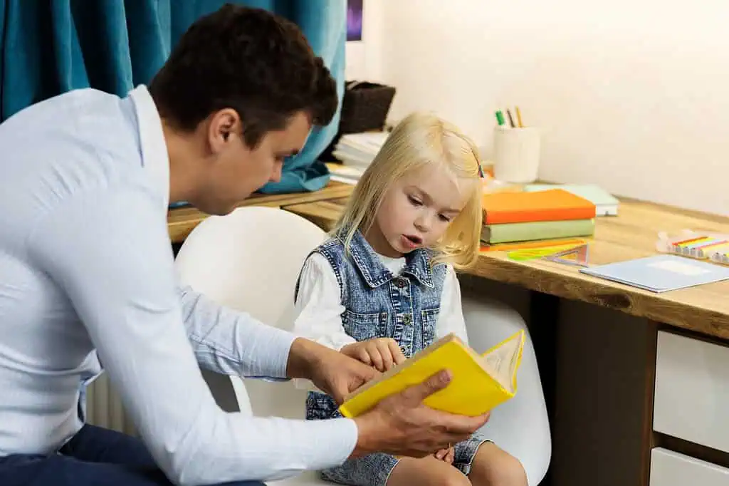 A young girl reads from a book with the guidance of an adult, symbolizing supportive learning strategies for children with Dyspraxia.