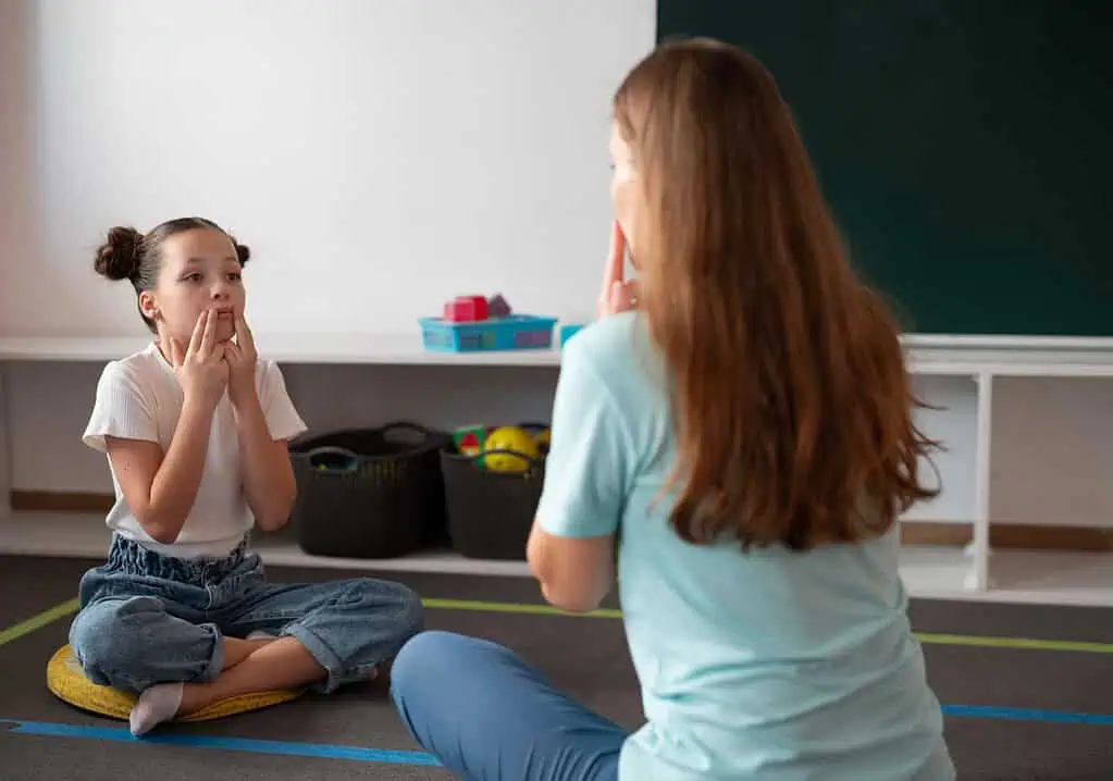 A young girl sits on the floor mimicking mouth movements during a speech therapy session with her instructor, illustrating supportive therapy for Dyspraxia.