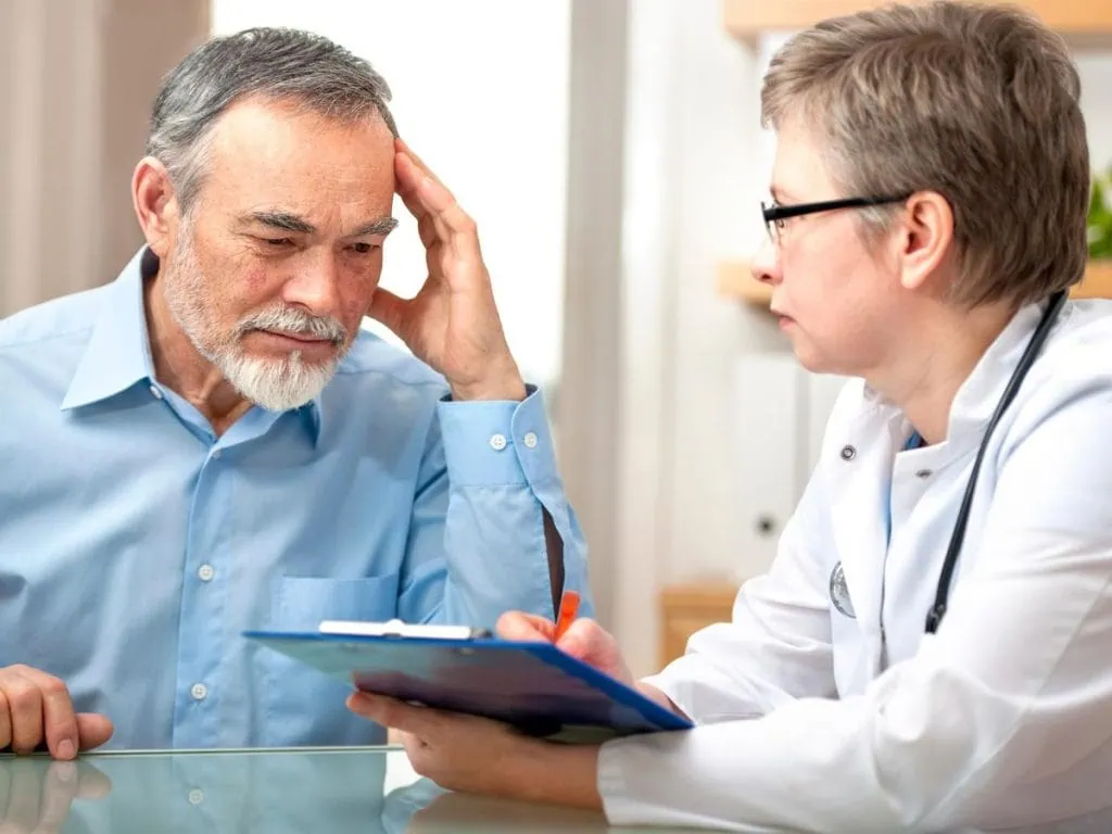 Concerned elderly man holding his head while speaking with a female doctor during a medical consultation, indicating stress or confusion.