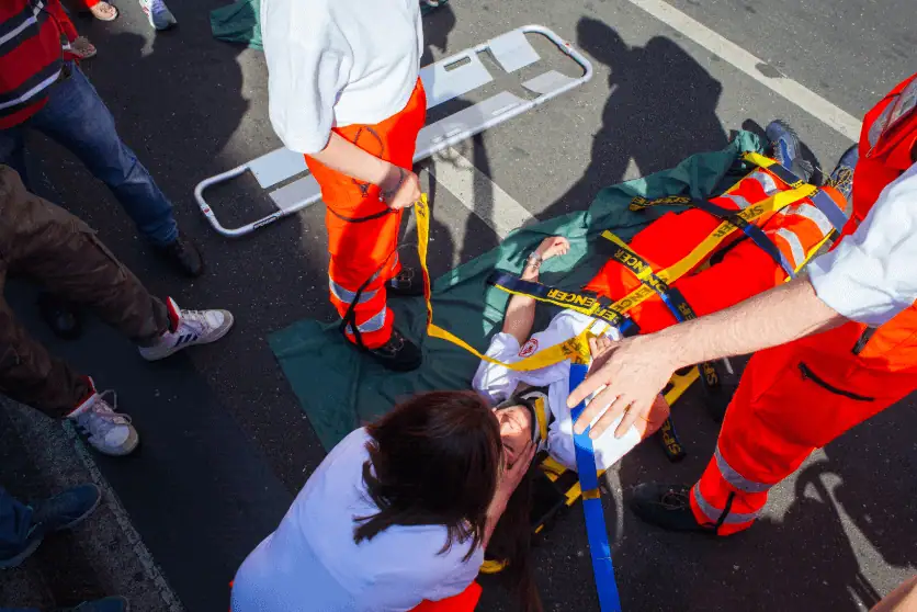 Emergency responders in orange and white uniforms attend to an injured person lying on a stretcher on a road. One paramedic secures the patient with yellow straps while another provides support. A concerned individual kneels beside the injured person, and bystanders observe the scene.