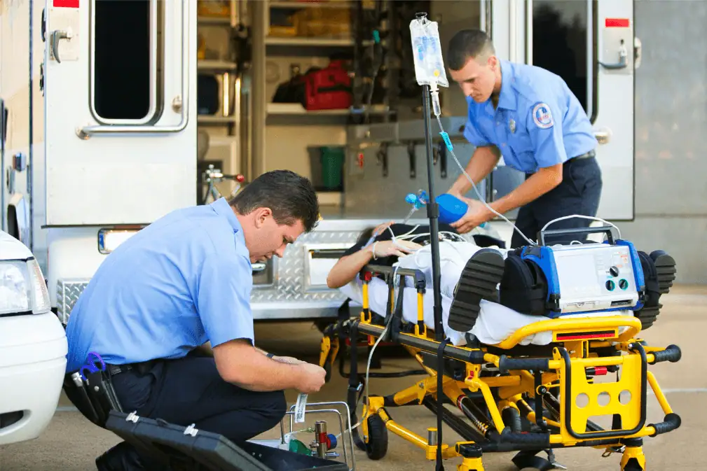 Two emergency medical responders providing life-saving treatment to a patient on a stretcher outside an ambulance. One paramedic is administering oxygen using a bag-valve mask, while the other prepares medical equipment.