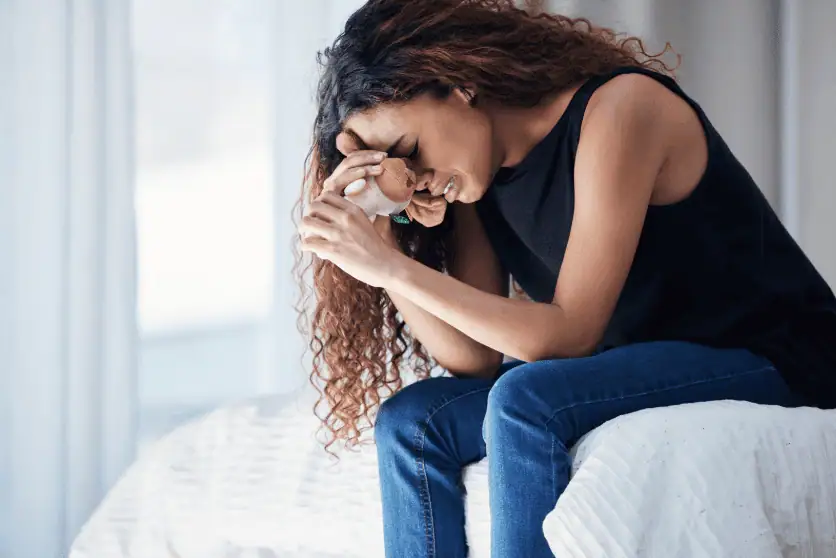 A distressed woman with curly hair sits on a bed, holding a small stuffed toy tightly against her forehead while crying. She wears a black sleeveless top and blue jeans, with her body slightly hunched forward. The background is softly lit, showing a white bedspread and curtains.