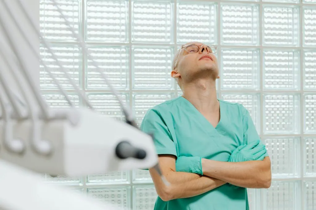 A healthcare worker in scrubs and a face mask sits with their head bowed and eyes closed, showing signs of exhaustion and emotional fatigue, reflecting the toll of demanding medical duties.