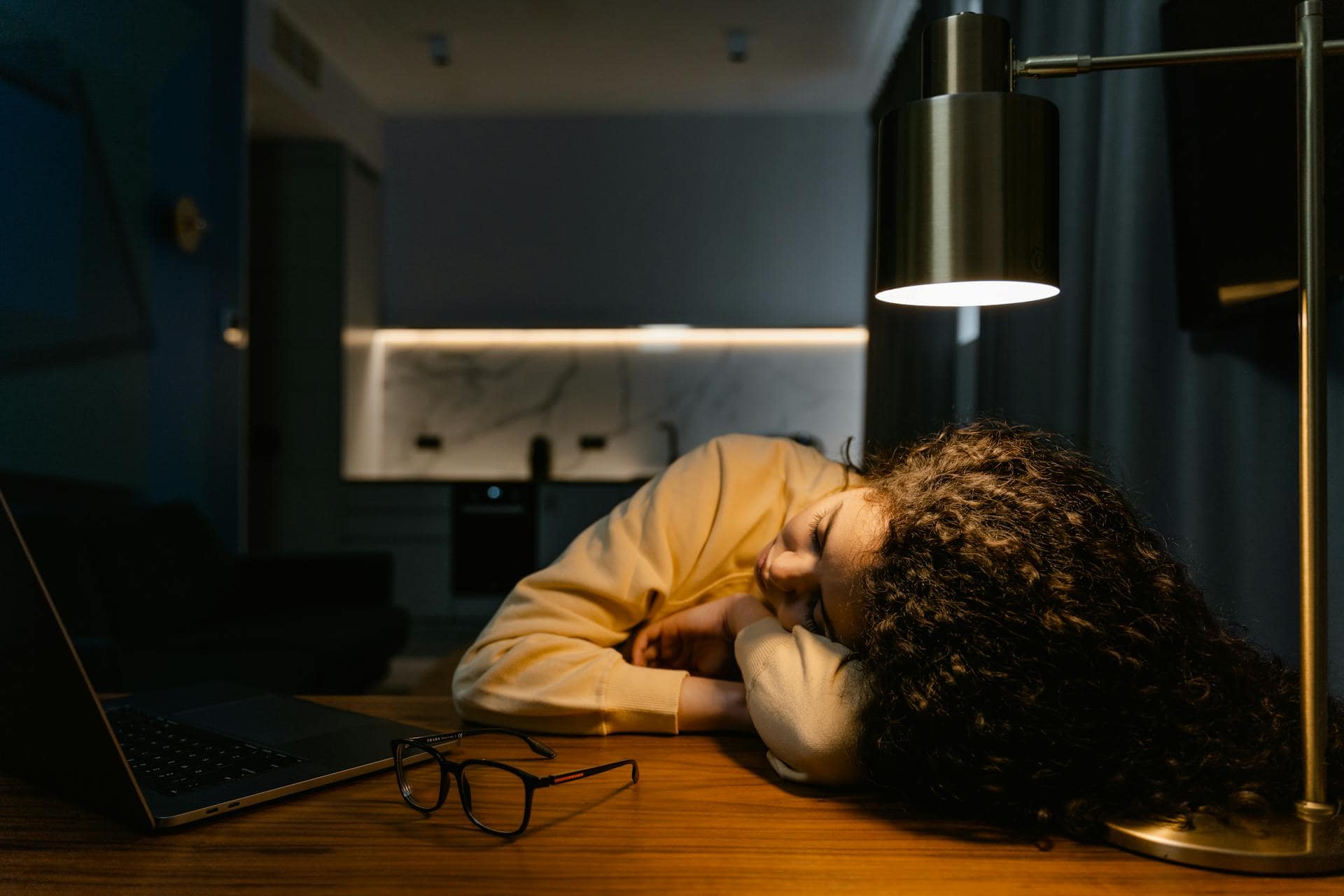 A girl with long curly hair rests her head on her arms on a desk under a lamp, beside a laptop and glasses