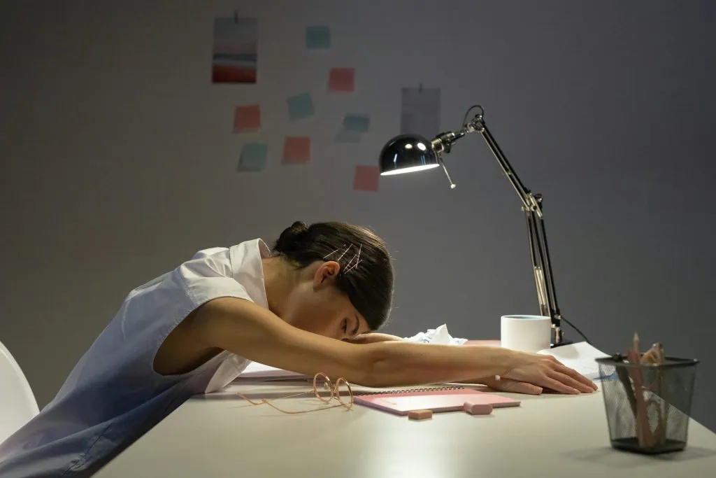A young woman in a white sleeveless blouse falls asleep at her desk, resting her head on her arms. A desk lamp illuminates her workspace, which includes a notebook, glasses, and scattered stationery. The background has sticky notes on the wall, suggesting a late-night work or study session.